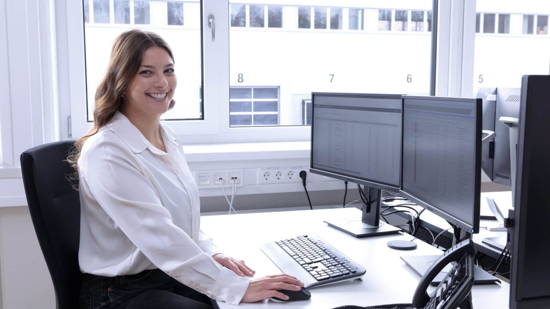 Frau in weißer Bluse sitzt an einem Bürotisch, lächelt in die Kamera, die Hände auf der Tastatur, vor zwei Computermonitoren, auf denen Tabellenkalkulationen angezeigt werden.